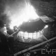 widok na płonącą kopułę oraz wypalone wnętrza budynku; widoczny strażacki wóz bojowy oraz strumień wody, którą strażacy próbują zgasić pożar; w oddali kamienice mieszkalne