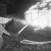 strażak w ubraniu bojowym w czasie akcji gaśniczej, polewający wodą z pompy płonący budynek; w tle krzaki, prowizoryczny płot z blachy falistej; w oddali wieżowce mieszkalne 