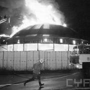 płonący budynek otoczony prowizorycznym płotem, przed którym stoi strażacki wóz bojowy; widoczny strażak biegnący w ubraniu bojowym oraz podnośnik z dwom strażakami gaszącymi ogień