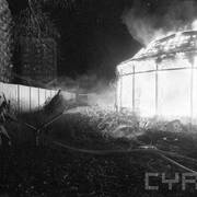 strażak w ubraniu bojowym w czasie akcji gaśniczej, polewający wodą z pompy płonący budynek; w tle krzaki, prowizoryczny płot z blachy falistej; w oddali wieżowce mieszkalne 