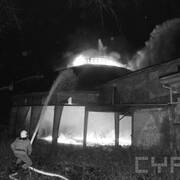 dwóch strażaków w ubraniach bojowych, jeden z nich trzyma wąż z wodą, którą polewa płonący budynek