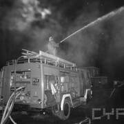 strażak w ubraniu bojowym stojący na dachu wozu strażackiego i polewający wodą z węża płonący budynek; w oddali widać innych strażaków gaszących pożar oraz fragmenty budynków mieszkalnych