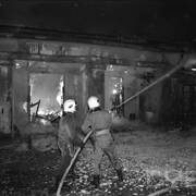 dwóch strażaków w ubraniach bojowych, trzymający wąż z wodą, którą polewają płonący budynek