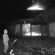 strażak w ubraniu bojowym, stojący przy pompie wodnej na tle płonącego budynku