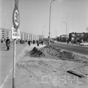 ulica z rozkopanym pasem ziemi wzdłuż jezdni, w głębi przechodnie i niskie bloki po lewej oraz wille po prawej stronie ulicy