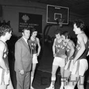 grupa mężczyzn w strojach sportowych stojąca wokół i słuchająca mężczyzny ubranego w garnitur; w głębi widać kosz do gry w koszyków i transparenty na ścianach