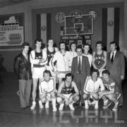 grupa mężczyzn, czterech kuca, reszta stoi; trzech mężczyzn nosi garnitury, a pozostali stroje sportowe; mężczyźni pozują do fotografii na boisku do koszykówki