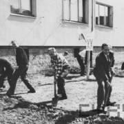 grupa osób przekopująca ziemię łopatami na tle budynku mieszkalnego z cegły