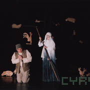 z lewej aktor w kostiumie klęczy ze złożonymi do modlitwy dłońmi, z prawej aktorka w kostiumie śmierci stoi nad nim z wyciągnięta kosą, dookoła z nich drewniane lalki zwierząt