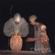 z lewej mężczyzna trzyma w rękach drewnianą lalkę, którą wyciąga z drewnianej małej beczki, z prawej kobieta wyciąga do nich ręce, przed nią stoi drewniana kukła kobiety