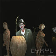 na pierwszym planie, po obu stronach zdjęcia drewniane lalki mężczyzny (z lewej) i kobiety, pośrodku drewniana beczka, w głębi mężczyzna w cylindrze i płaszczu oraz ze sznurem na szyi