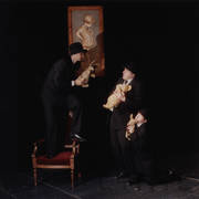 scena ze spektaklu, w której biorą udział dwaj mężczyźni i kobieta w czarnych garniturach i kapeluszach na głowach w rękach trzymają alki ze spektaklu, z lewej na fotelu stoi mężczyzna z lalką Królika, pośrodku mężczyzna z lalką misia, z prawej klęczy kobieta z lalką Prosiaczka, za nimi portret chłopca