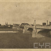 stalowy most na pięciu przęsłach i murowanych, masywnych filarach, na moście ozdobne latarnie, w głębi budynki, pośrodku kościół z dwiema wieżami