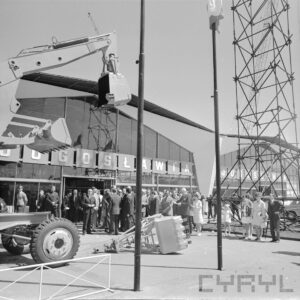 koparki na betonowy placu, w głębi grupa ludzi, za nimi przeszklony budynek