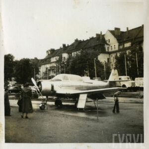 grupka osób przyglądająca się samolotowy stojącemu na placu na tle drzew i kamienicy