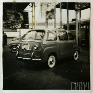 samochód stojący na placu targowym, w tle widoczne fragmenty innych samochodów i banery reklamowe