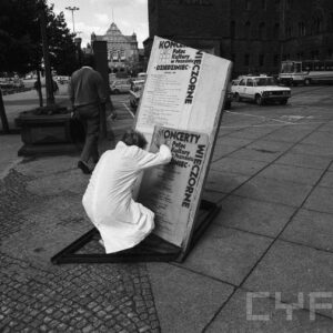 kobieta przyklejająca plakat na tablicę, na drugim planie budynek z kamienia i samochody