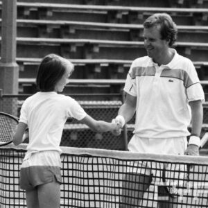 mężczyzna w średnim wieku w stroju sportowym, trzymający w dłoni rakietę tenisową gratulujący młodej dziewczynie ubranej w strój sportowy i również trzymającej rakietę tenisową; w tle trybuny wokół kortu tenisowego