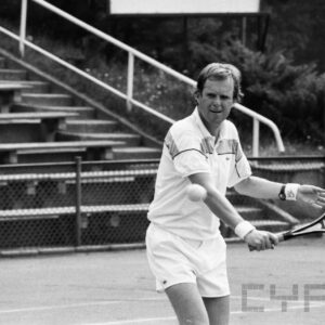 mężczyzna w średnim wieku w stroju sportowym, trzymający w dłoni rakietę tenisową i odbijający piłkę; w tle trybuny wokół kortu tenisowego