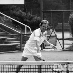 mężczyzna w średnim wieku w stroju sportowym, trzymający w dłoni rakietę tenisową i odbijający piłkę; w tle trybuny wokół kortu tenisowego