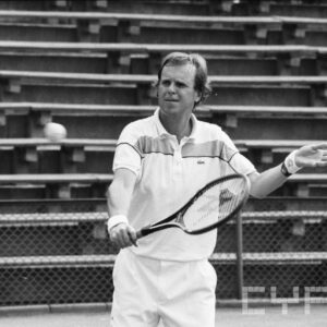 mężczyzna w średnim wieku w stroju sportowym, trzymający w dłoni rakietę tenisową i odbijający piłkę; w tle trybuny wokół kortu tenisowego