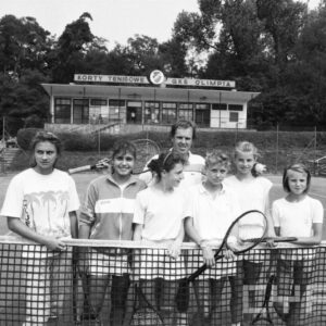 mężczyzna w średnim wieku z sześcioma dziewczętami, ubrani w stroje sportowe, stoją przy siatce na korcie tenisowym; w tle budynek klubowy