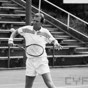 mężczyzna w średnim wieku w stroju sportowym, trzymający w dłoni rakietę tenisową i odbijający piłkę; w tle trybuny wokół kortu tenisowego