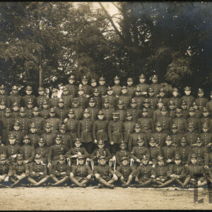 Grupa kilkudziesięciu mężczyzn w różnym wieku w mundurach wojskowych na tle drzew. Większość z nich stoi, część siedzi.
