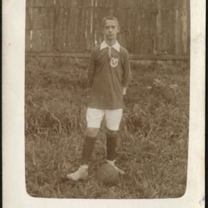 chłopiec ubrany w strój sportowy, pozujący z piłką na tle drewnianego płotu i trawy