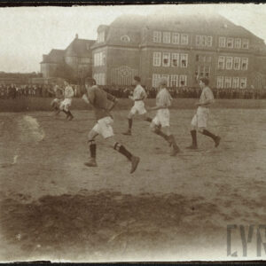 Grupa młodych mężczyzn w strojach sportowych grająca w piłkę nożną na boisku, w tle grupa ludzi obserwujących mecz oraz wysoki budynek z cegieł.