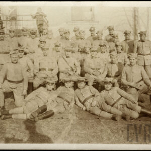 Grupa mężczyzn w różnym wieku w mundurach wojskowych na tle budynku. Większość z nich stoi, część siedzi, a kilku leży. Za mężczyznami stoi trójka dzieci.