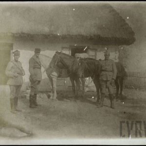 Trójka mężczyzn w mundurach wojskowych stojąca przy trzech koniach na tle chaty pokrytej strzechą.