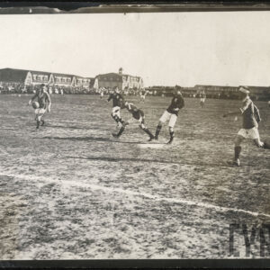 Grupa młodych mężczyzn w strojach sportowych grająca w piłkę nożną na boisku, w tle grupa ludzi obserwujących mecz oraz wysokie budynki z cegieł.