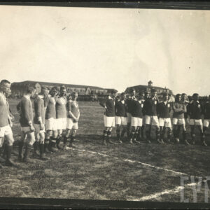 Dwie grupy młodych mężczyzn w strojach sportowych stojące na boisku do gry w piłkę nożną. Mężczyźni ubrani są w ciemne koszulki z białymi kołnierzami, jasne krótkie spodenki, ciemne getry i ciemne buty piłkarskie. W tle stoi grupa ludzi obserwujących mecz, widoczne są również budynki z cegieł.  