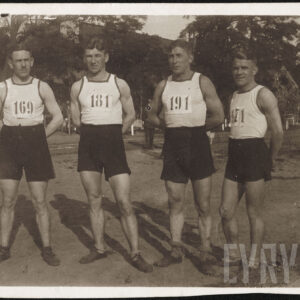 Czterech mężczyzn w strojach gimnastycznych, składających się z białych koszulek na ramiączkach i ciemnych krótkich spodenek, stojących na tle drzew. Na koszulkach widoczne numery startowe.