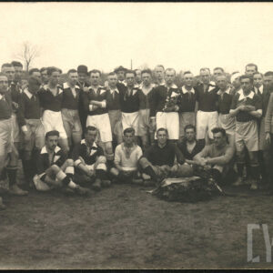 Grupa mężczyzn w strojach sportowych, kilku z nich nosi garnitury. Część mężczyzn stoi, kilku z nich siedzi. Przed siedzącymi mężczyznami leży piłka do gry otoczona wieńcem z liści z pamiątkową szarfą.
