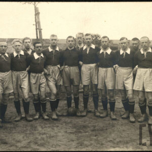 Grupa młodych mężczyzn stojących w strojach sportowych składających się z ciemnych koszulek z białym kołnierzem, jasnych krótkich spodenek, czarnych getrów i ciemnych butów piłkarskich. Przed jednym z mężczyzn leży piłka do gry. 