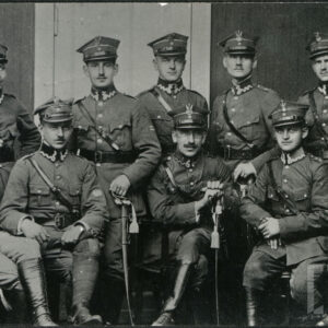 Grupa mężczyzn w różnym wieku w wojskowych mundurach oficerskich wewnątrz budynku. Kliku z nich stoi, kilku siedzi. W tle na ścianie wisi ramka ze zdjęciem psa.