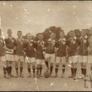 Grupa chłopców w strojach sportowych stojąca na boisku do gry w piłkę nożną na tle drzew. Chłopcy ubrani są w ciemne koszulki z białym kołnierzem, jasne krótkie spodenki, czarne getry i ciemne buty piłkarskie. Jeden z chłopców trzyma stopę na piłce do gry. 