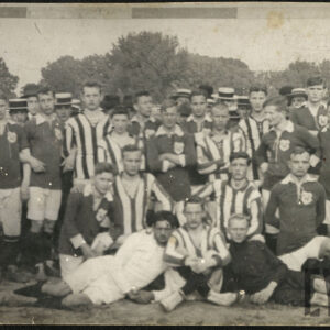 Grupa młodych mężczyzn w strojach sportowych, kilkoro w kapeluszach z rondem, stoi, klęczy lub leży na tle drzew.