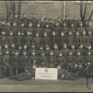 Grupa mężczyzn w różnym wieku w mundurach wojskowych, na tle ceglanego budynku i drzew. Większość z nich stoi, kilku siedzi, a dwóch leży. Pomiędzy leżącymi stoi karton z saperskim znakiem graficznym i nazwą kompanii.