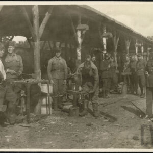 Grupa mężczyzn w różnym wieku, w mundurach wojskowych, stojąca przy stanowiskach w prowizorycznej kuźni. Jeden z mężczyzn formuje podkowę na kowadle.