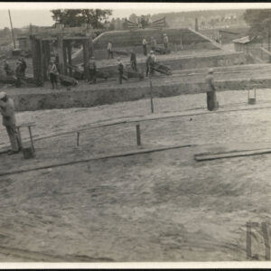 Grupa mężczyzn pracująca przy budowie nasypów i mostu. Część z nich trzyma w rękach taczki. W tle drzewa i zabudowania drewniane. 