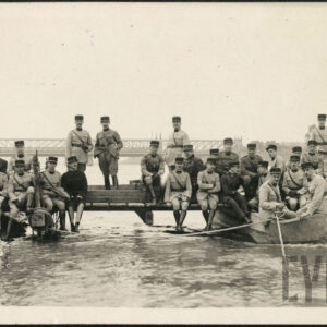 Grupa kilkudziesięciu mężczyzn w różnym wieku we francuskich mundurach wojskowych. Trzech z nich stoi, pozostali siedzą na drewnianym moście, część siedzi w łodziach przycumowanych do mostu. W oddali most kolejowy.