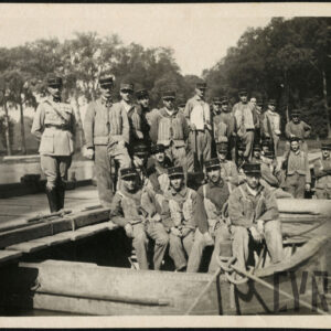 Grupa kilkudziesięciu mężczyzn w różnym wieku we francuskich mundurach wojskowych i w kapokach. Część z nich stoi na drewnianym moście, pozostali siedzą w łodziach przycumowanych do mostu. W tle drzewa.