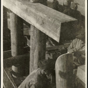 Grupa mężczyzn w roboczych strojach wojskowych pracująca przy budowie drewnianego mostu. Jeden z nich dokonuje pomiaru przy pomocy poziomicy. 