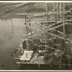 Grupa mężczyzn w roboczych strojach wojskowych pracująca przy budowie drewnianego mostu nad akwenem.