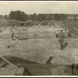 Mężczyzna w wojskowym mundurze oficerskim stojący na nasypie ziemnym. W tle mężczyźni pracujący przy budowie nasypu i mostu. Na drugim planie drzewa i murowane zabudowania cywilne. 