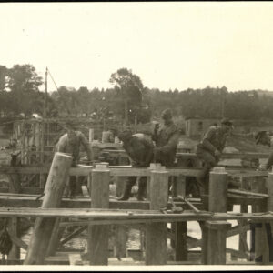 Grupa mężczyzn w roboczych strojach wojskowych pracująca przy budowie drewnianego mostu na tle drzew.