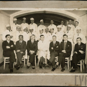 duża grupa mężczyzn, kilkoro w garniturach (jeden w kitlu) siedzi na krzesłach, pozostali ubrani w białe kitle i czapki stoją